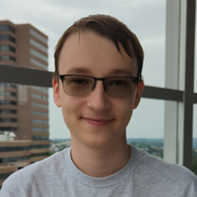 Portrait of me with glasses in a light grey shirt with a window in the background. A brick and glass building is visible through the window on the left side of the image.